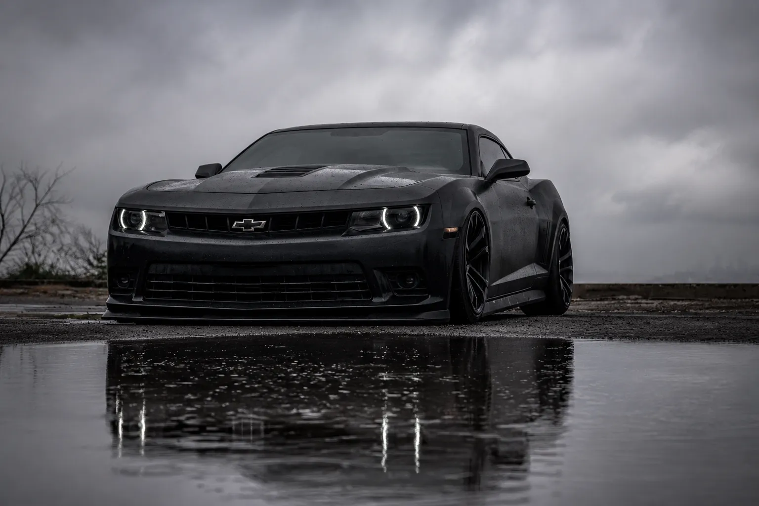 Black Chevrolet Camaro parked on wet pavement under a cloudy sky, showcasing a sleek performance car often associated with professional Chevrolet service & maintenance styling and care.