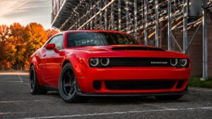A bright red Dodge muscle car is parked in an outdoor industrial setting, showcasing its bold front grille, signature Dodge badge, aggressive stance, and wide-body styling. Captured during golden hour with autumn trees in the background, the image reflects the performance and appearance maintained by a trusted Dodge repair center offering expert service and maintenance.